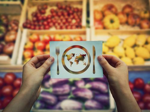 hands holding an image of a knife, fork and plate with the the world inside, in front of a fruit and vegetable market stall