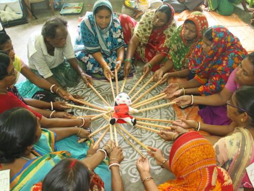 a circle of seated figures hold out sticks supporting a doll