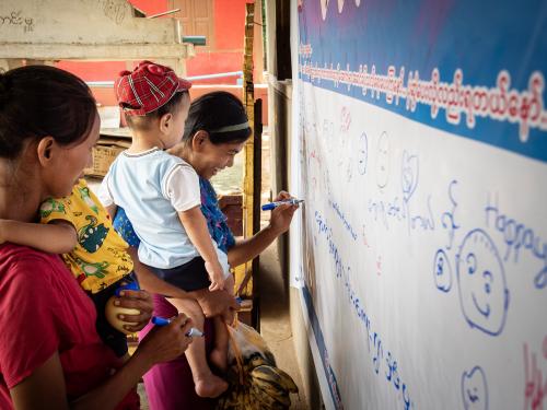 Two women while carrying their young children write on a big white banner
