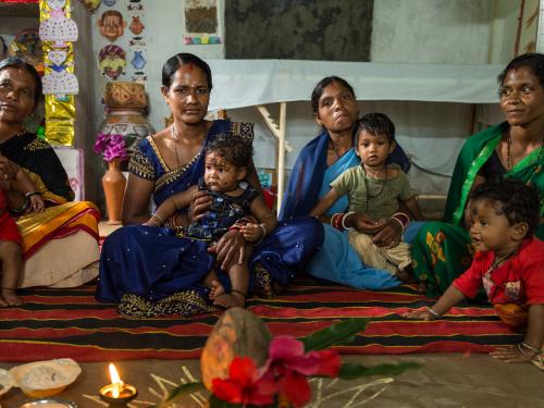 Four women sit on the floor with their young children with four bowls of complementary food, a lit oillamp and a decorated coconut in front of them