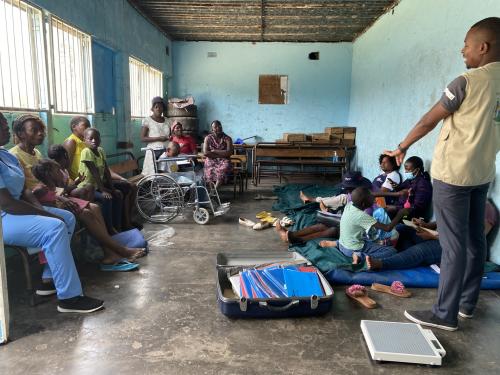 A group of women with their children sit around a room listening to a man standing and talking. In the foreground there is an open suitcase with multiple files and a weighing scale