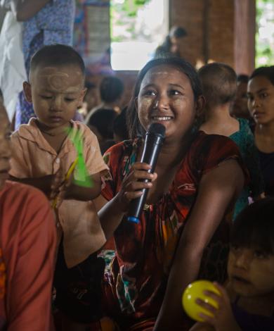 A room full of women with their children focuses on one woman who holds a microphone as she speaks.