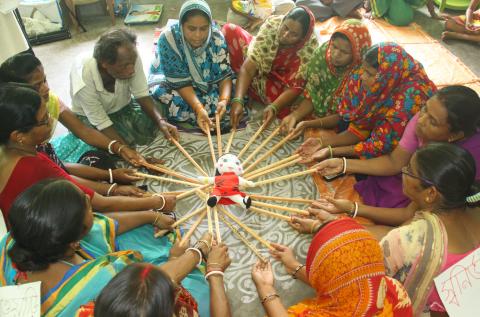 a circle of seated figures hold out sticks supporting a doll