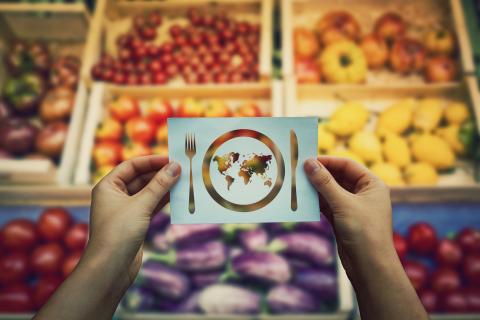 hands holding an image of a knife, fork and plate with the the world inside, in front of a fruit and vegetable market stall
