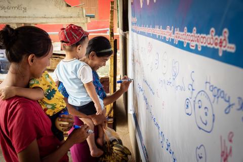 Two women while carrying their young children write on a big white banner