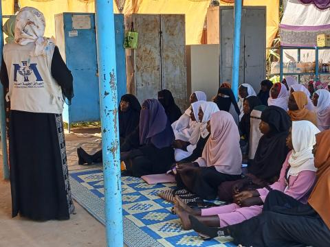 A group of women with their young children sitting in a row while a women stands and speaks to them