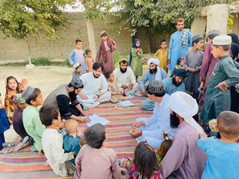 A group of men and children sit and stand outside on a floor mat while looking on to one man who is taking notes. A MUAC tape can also be seen. 